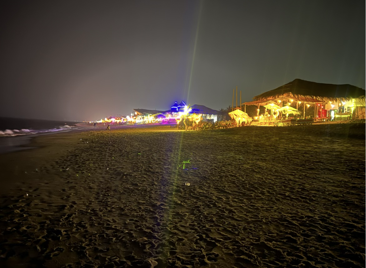 A Goa beach at night, a long strip of shacks lit by coloured LED rope-light, the sand glowing orange for hundreds of metres