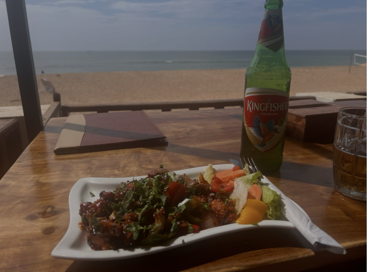 A plate of chilli chicken and a cold beer on a wooden table, the beach and sea just beyond